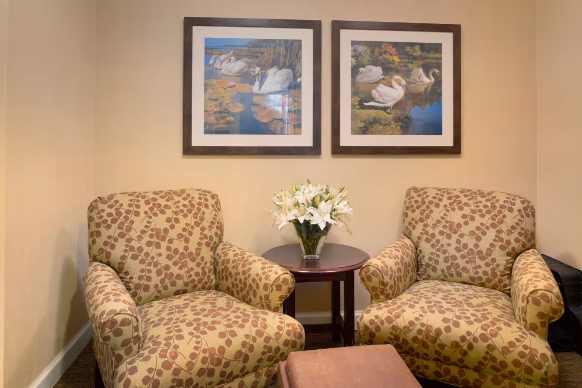 Two patterned armchairs with a small round wooden table between them holding a vase of white flowers, set against a beige wall with two framed pictures of swans above.