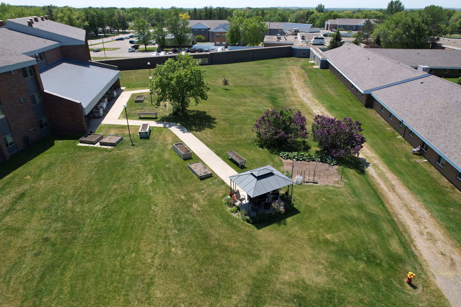 Aerial view of a senior living community outdoor area with green lawns, a paved walkway, benches, raised garden beds, a small gazebo, and flowering bushes. Surrounding the lawn are brick buildings with multiple windows and a parking lot in the background.