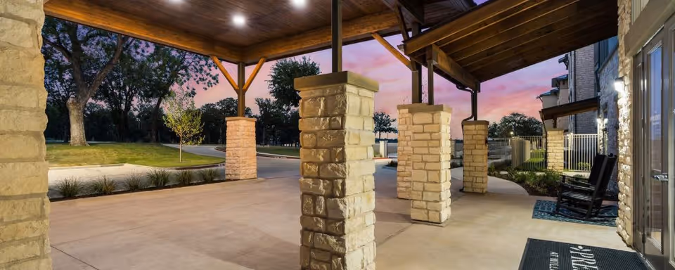 Covered stone-pillared entrance with a wooden ceiling, rocking chairs, and walkway at dusk.