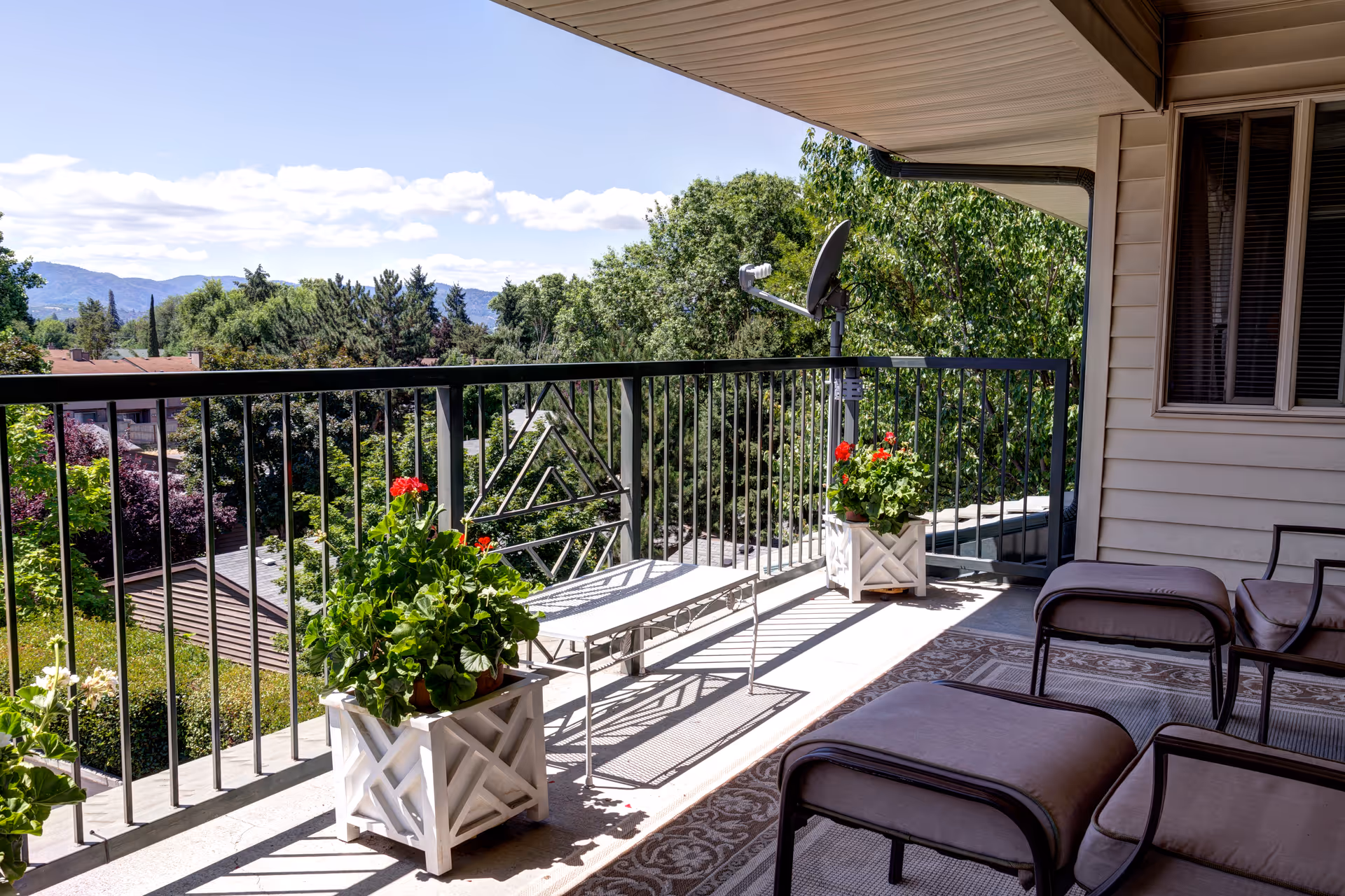 Covered balcony with lounge chairs, potted flowers, and a railing overlooking trees and distant hills.