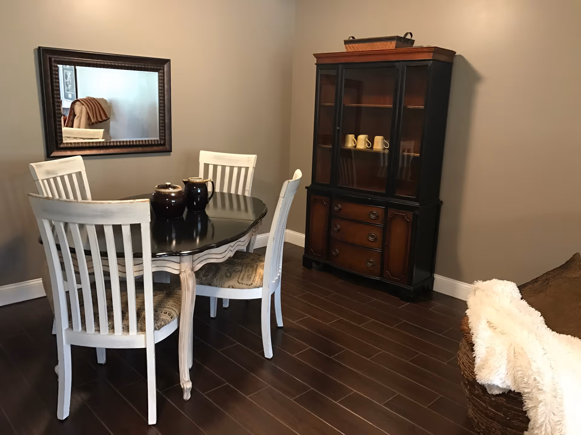 A dining area with a black oval table and four white wooden chairs with patterned cushions. On the table are two dark-colored ceramic pitchers. A large mirror with a dark frame hangs on the beige wall behind the table. To the right, there is a dark wooden cabinet with glass doors displaying several mugs. The floor is dark wood, and a wicker basket with a white fluffy blanket is partially visible in the corner.