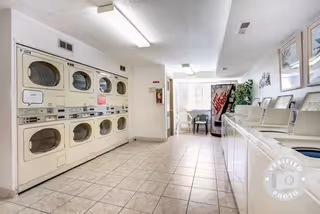 Laundry room with stacked washers and dryers on the left side and top-loading washing machines on the right side. The room has tiled flooring, fluorescent ceiling lights, a vending machine and a small seating area near the back window.