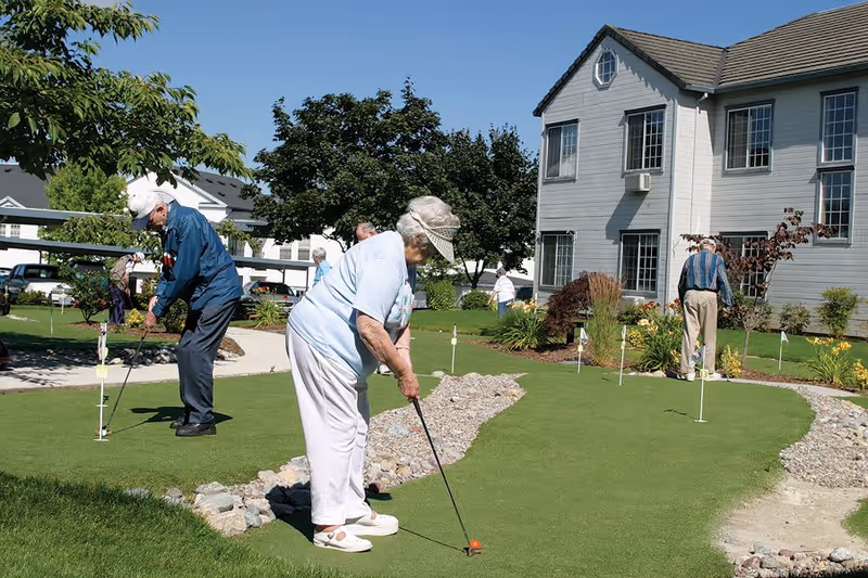 Several elderly residents play putt-putt on a putting green outside a senior living building on a sunny day.