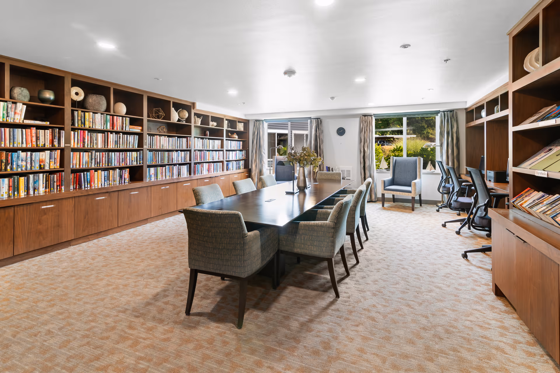 A well-lit room with a long wooden table surrounded by upholstered chairs, flanked by large bookshelves filled with books and decorative items. There are windows with curtains letting in natural light, and a few office chairs and desks along the right side of the room.