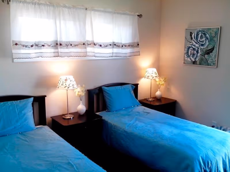 A bedroom with two single beds each covered with blue bedding and matching pillows. Between the beds is a dark wooden nightstand with a floral-patterned lamp and a small white vase with flowers. A window with white curtains is above the nightstand, and a framed painting of blue flowers hangs on the adjacent wall.
