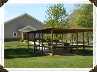 Outdoor covered picnic area with tables and benches on a well-maintained grassy lawn, adjacent to a beige building with a gabled roof and surrounded by trees and shrubs.