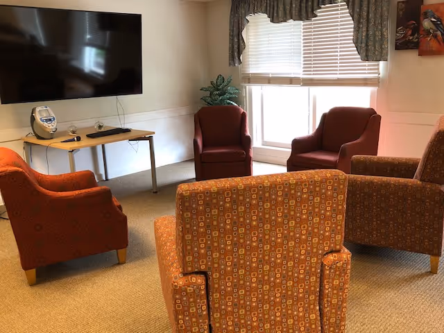 A small common room with upholstered armchairs arranged facing a wall-mounted TV and a window with blinds.