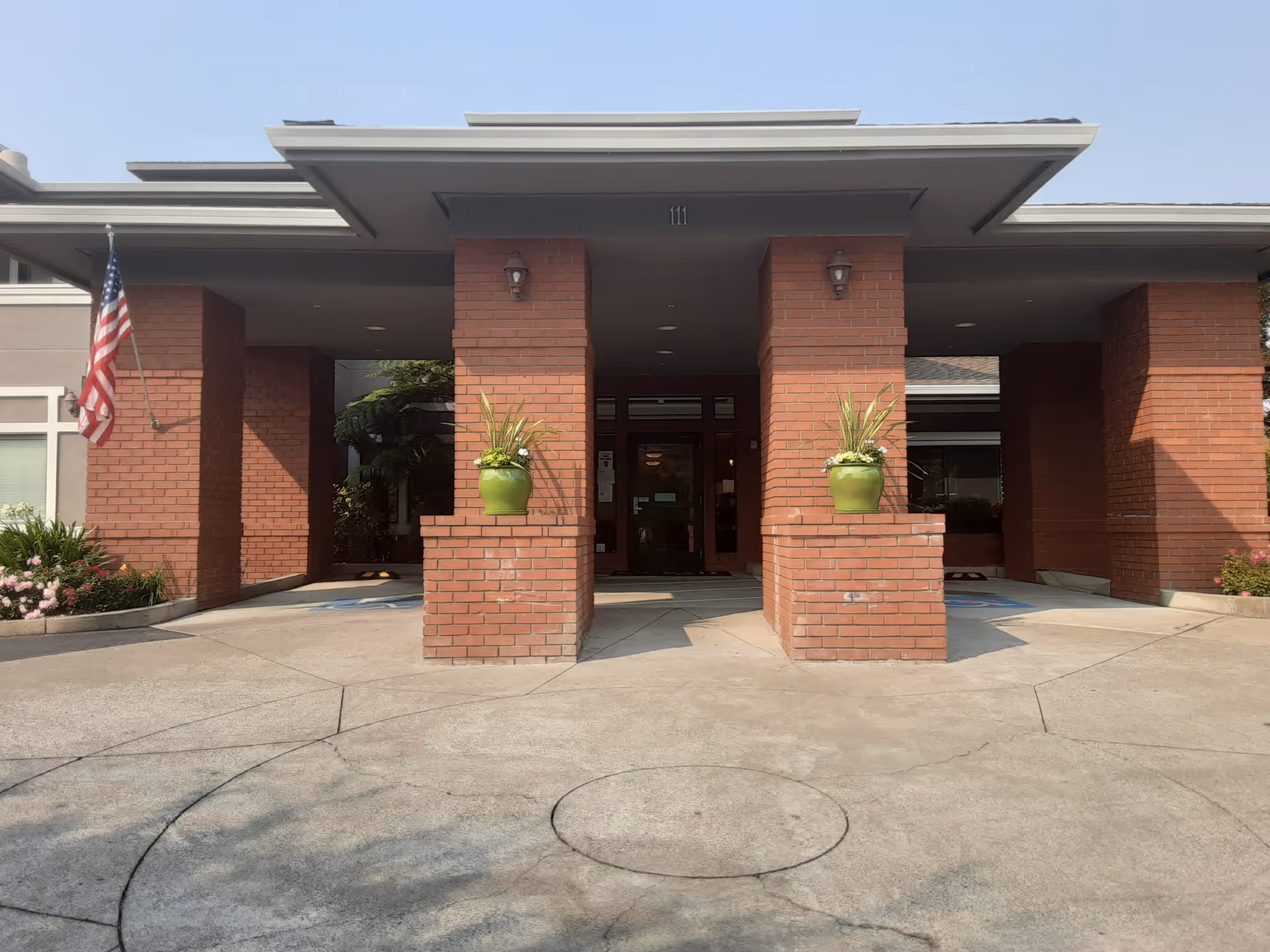 Front entrance of a brick building with two large brick pillars, each holding a green planter with plants. An American flag is mounted on the left side. The entrance has a covered driveway and glass doors leading inside.