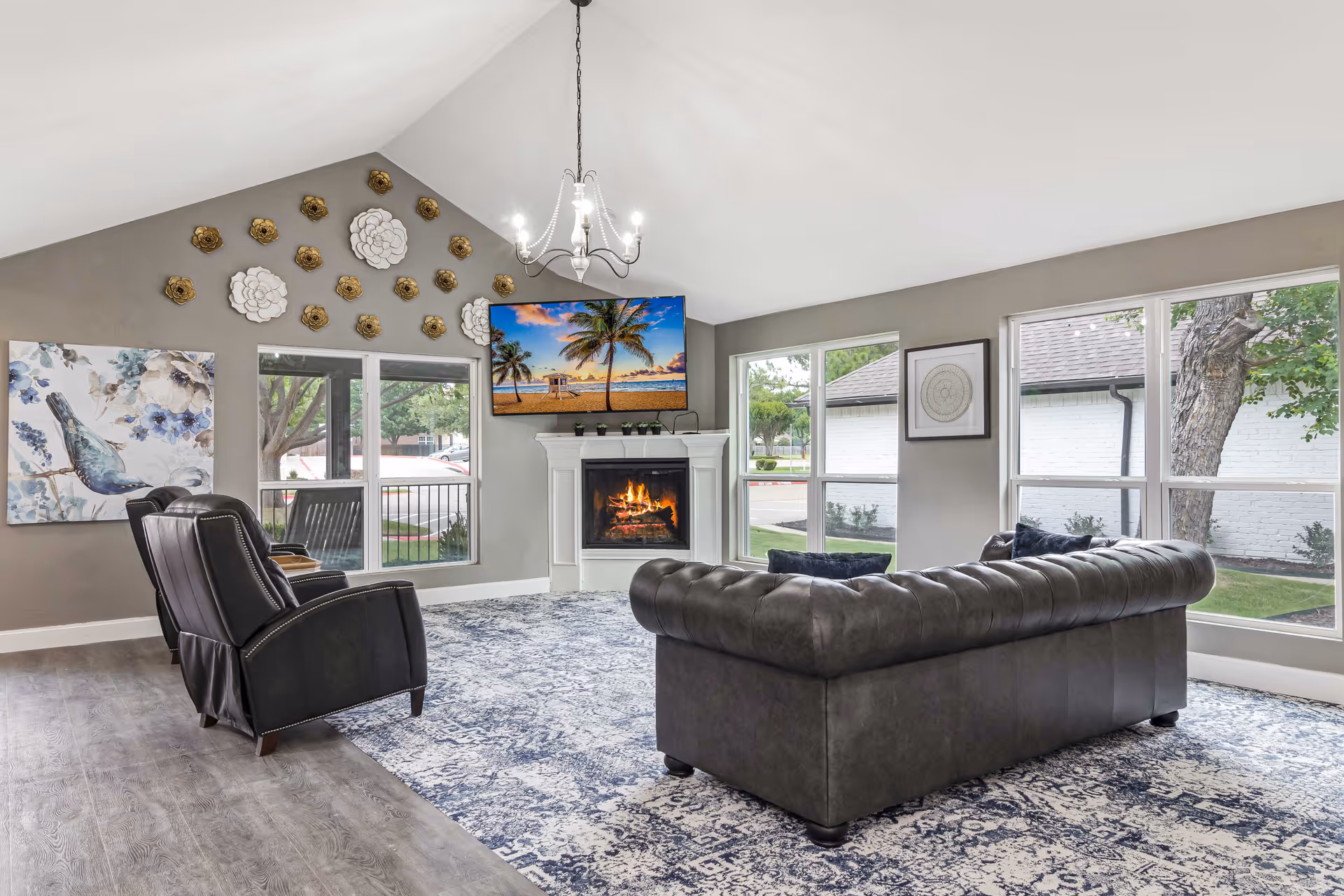 A bright living room with a vaulted ceiling, featuring a dark gray leather sofa and two black armchairs arranged around a fireplace with a TV mounted above it displaying a tropical beach scene. The walls are painted gray and decorated with floral wall art and a large painting of a bird. Large windows let in natural light and show an outdoor view with trees and a white building.