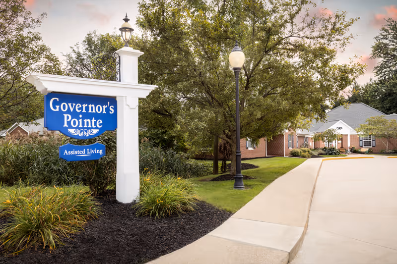 A blue 'Governor's Pointe Assisted Living' sign by a landscaped driveway leading to brick assisted-living buildings and a lamp post.