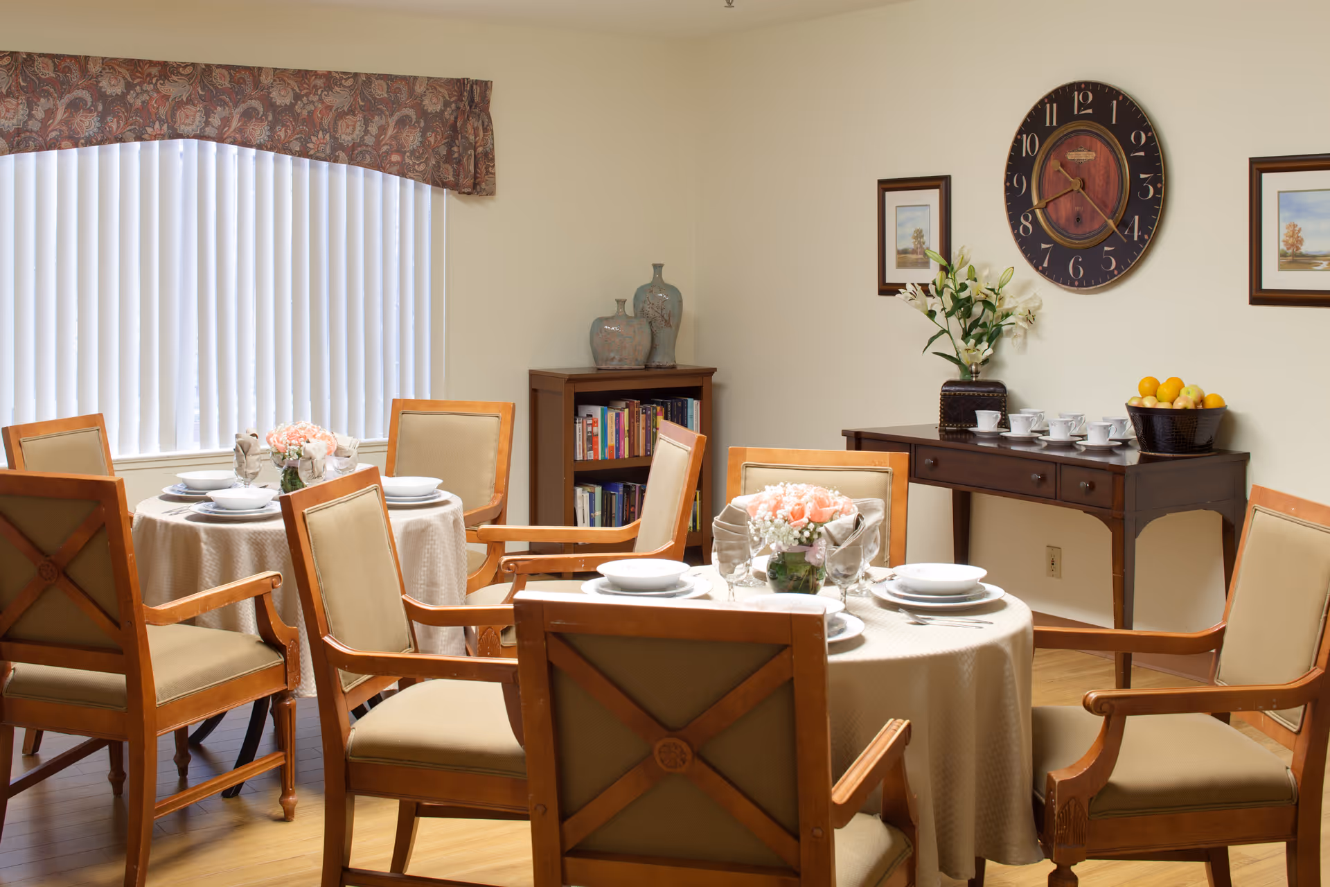 A dining room with two round tables covered with beige tablecloths, each set with white plates, bowls, and napkins. There are six wooden chairs with beige cushions around the tables. A window with vertical blinds and a patterned valance is on the left wall. Against the back wall, there is a wooden sideboard with a basket of fruit, a vase with flowers, and several cups and saucers. Above the sideboard, a large round clock and two framed pictures hang on the wall. A small bookshelf with decorative vases is in the corner.