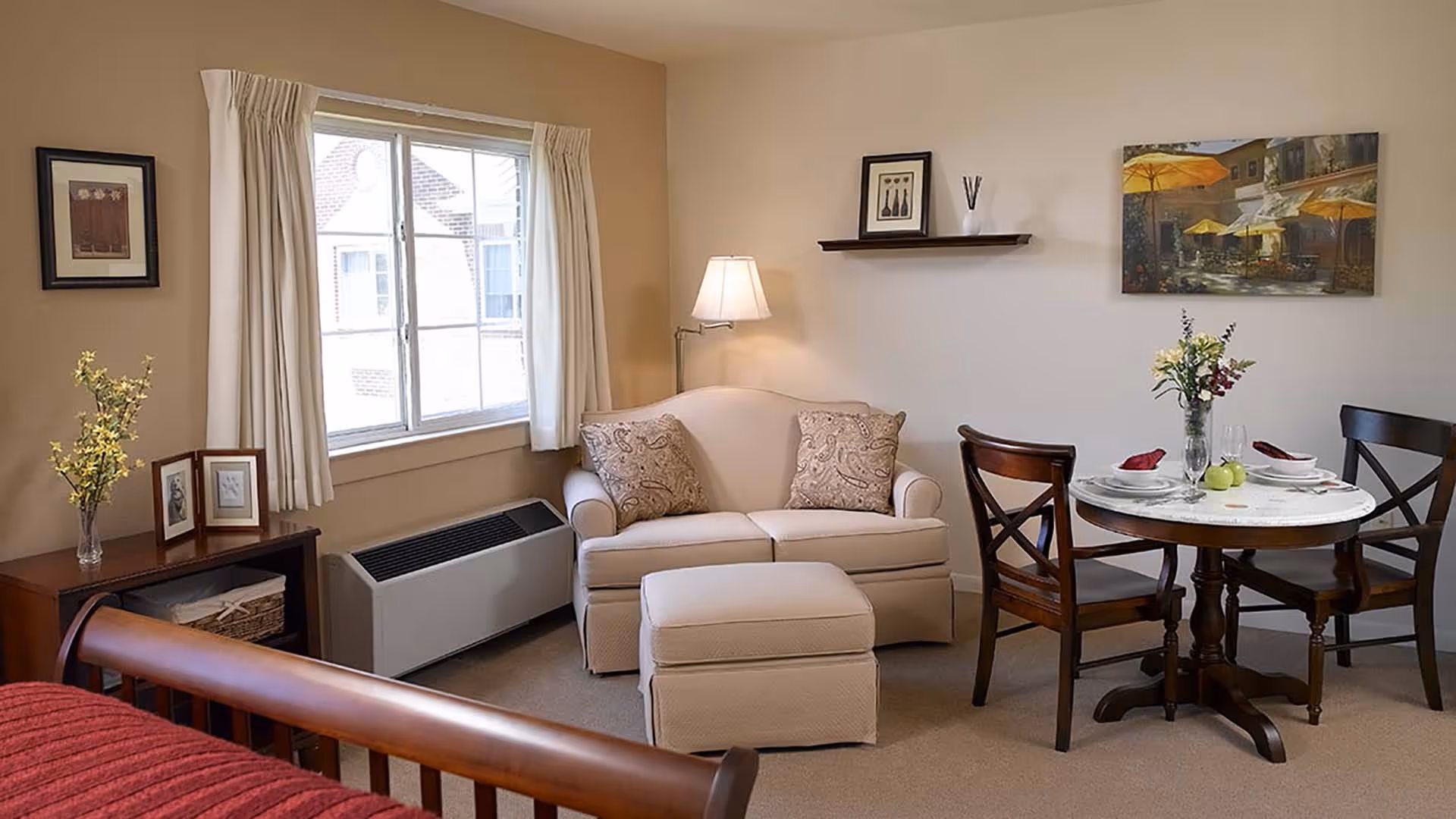 A cozy living area in a senior living facility featuring a beige loveseat with two patterned pillows, a matching ottoman, a round wooden dining table set for two with flowers and napkins, a wooden side table with framed photos and a vase of flowers, a window with cream curtains, and wall art including a painting and a framed picture on a floating shelf.