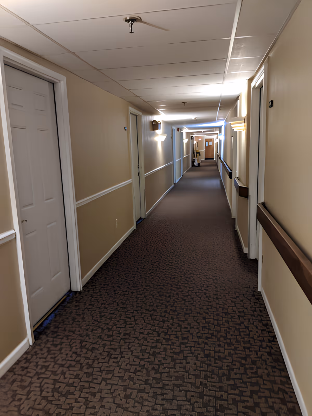 A long indoor hallway with beige walls and patterned brown carpet. Several white doors line both sides of the hallway, with handrails along the walls. Ceiling lights and wall sconces illuminate the corridor.