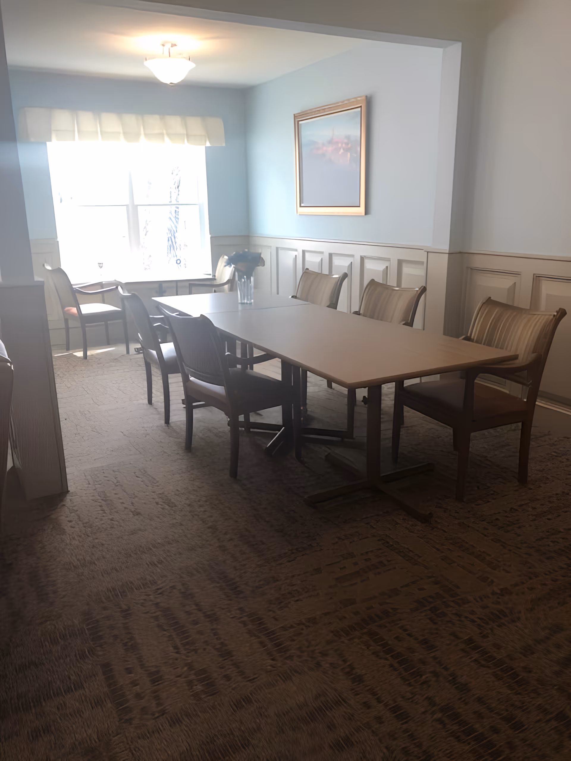 A dining room with a rectangular table surrounded by eight wooden chairs with cushioned seats. The room has light blue walls with white wainscoting, a window with a white valance allowing natural light to enter, a ceiling light fixture, and a framed painting on the wall. A vase with flowers is placed on the table.