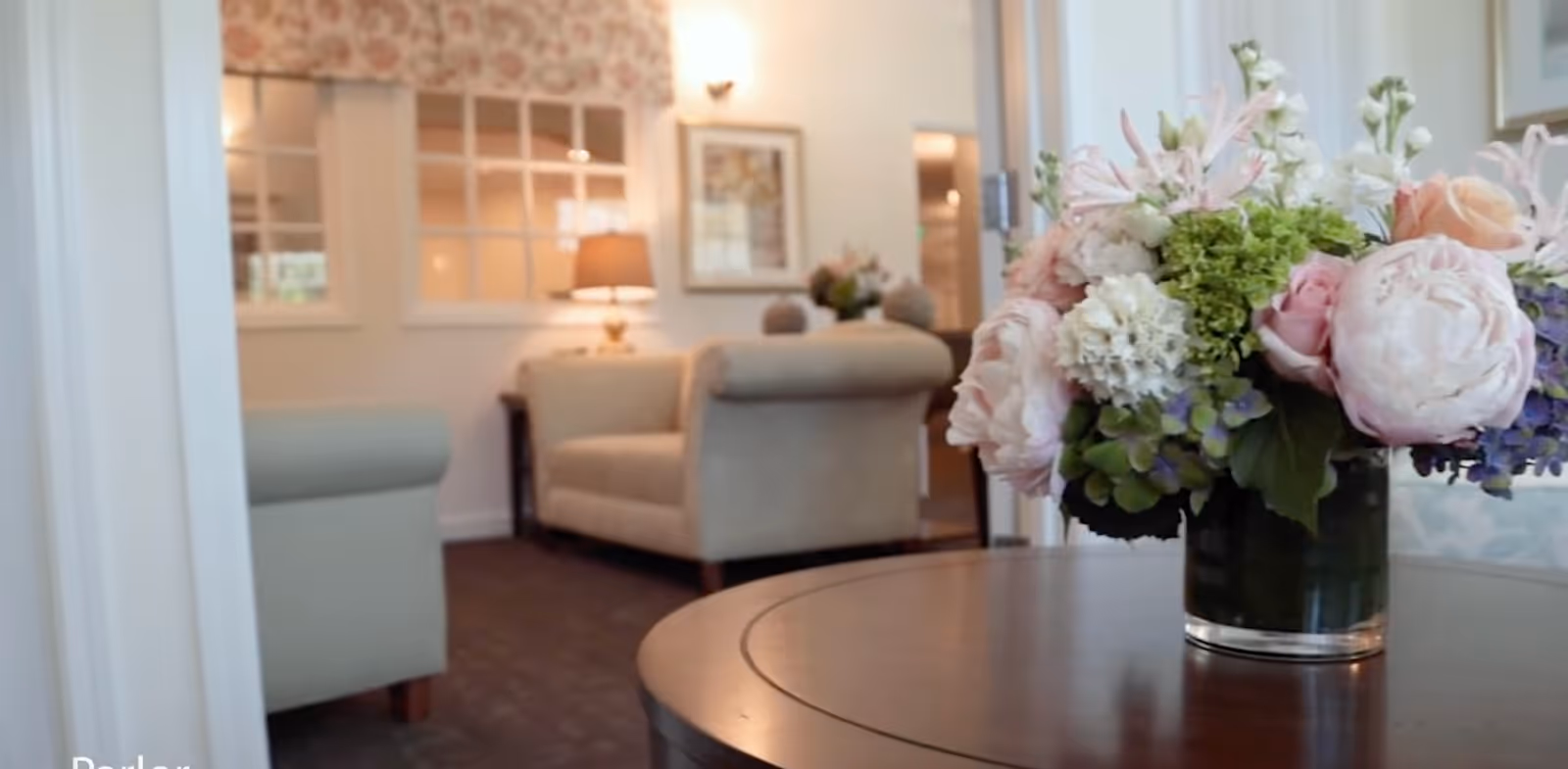 A cozy living room area with beige upholstered armchairs, a table with a floral arrangement in the foreground, a lamp on a side table, and windows with floral valances in the background.
