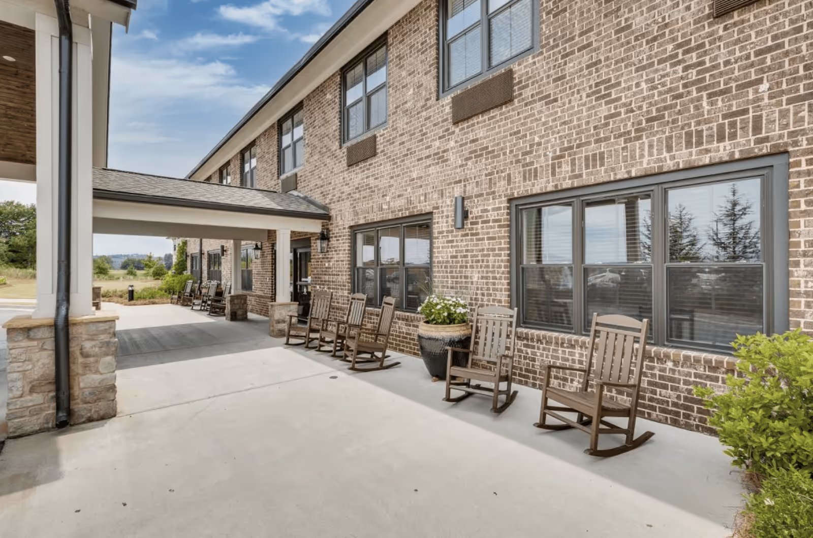 Outdoor covered patio area of a senior living facility with several wooden rocking chairs lined up against a brick wall under a roof extension. There are large windows on the building and potted plants placed near the chairs. The sky is partly cloudy.