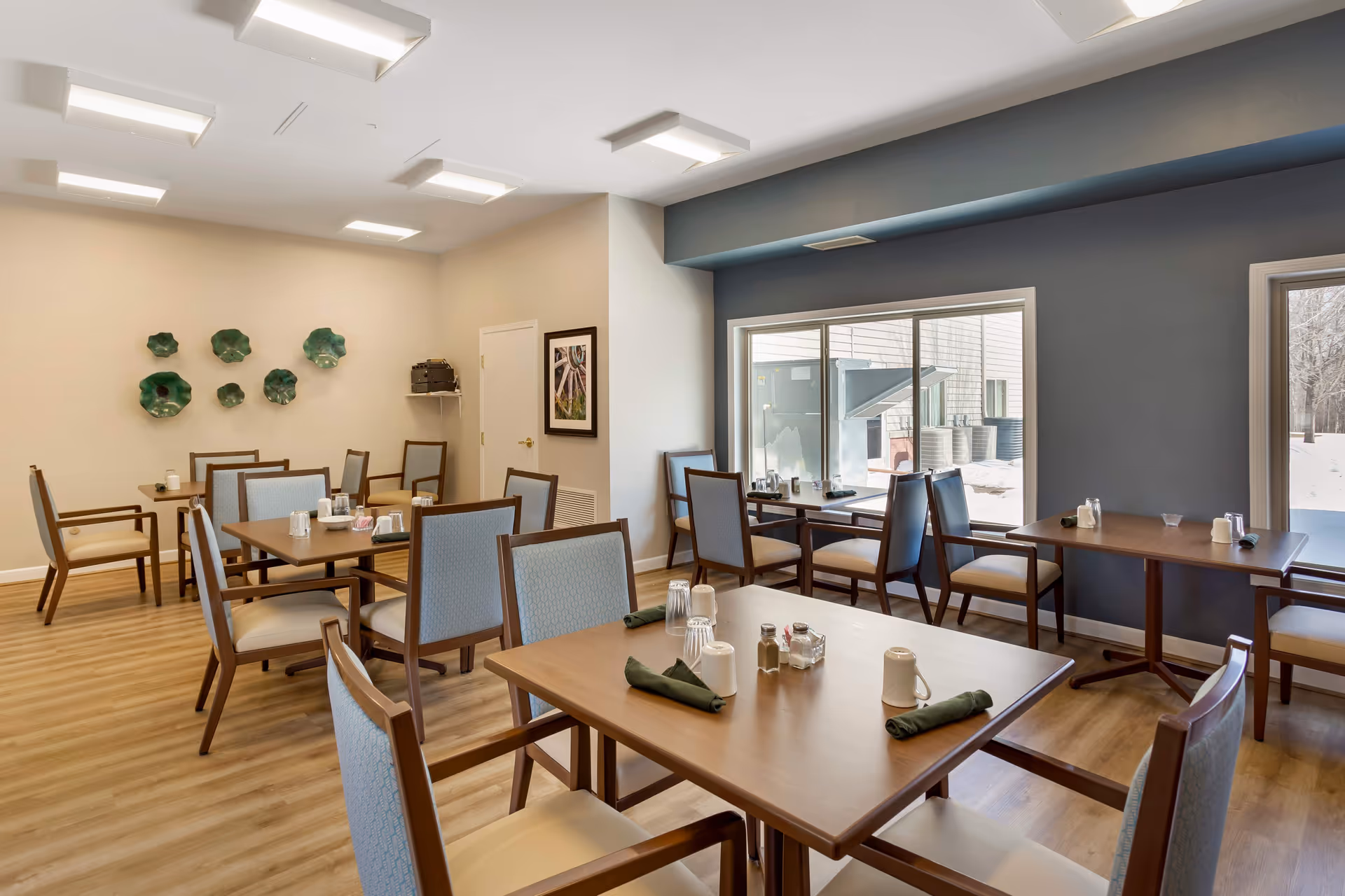 Bright dining room with multiple wooden tables and chairs set with napkins and condiments, large windows and decorative wall art.