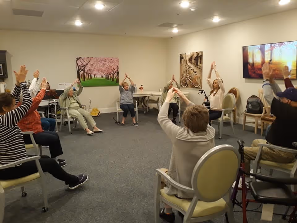 A group of elderly people seated in a circle in a room, participating in a seated exercise or stretching activity with their arms raised above their heads. The room has neutral-colored walls with three large framed pictures, and chairs arranged in a circle.