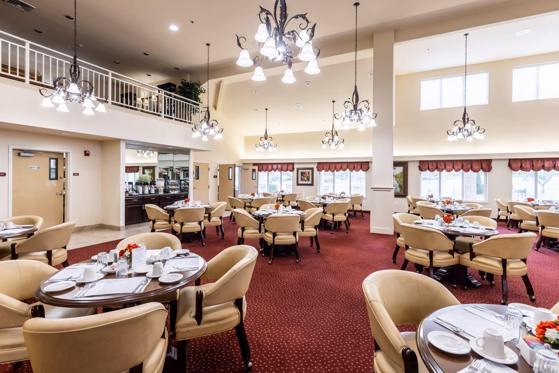 A spacious dining room in a senior living facility with multiple round tables set with white plates, cups, silverware, and small floral centerpieces. The room has beige walls, large windows with red valances, red carpet flooring, and several ornate chandeliers hanging from a high ceiling. There is a balcony area above and a buffet station along one wall.