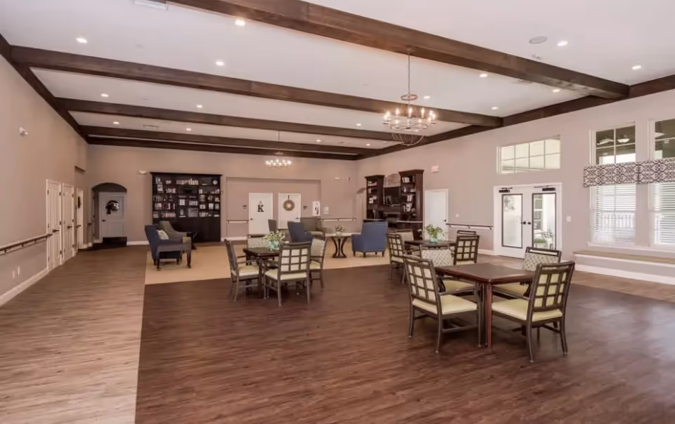 Spacious senior living common area with dining tables and chairs, lounge seating, bookshelves, and exposed ceiling beams.