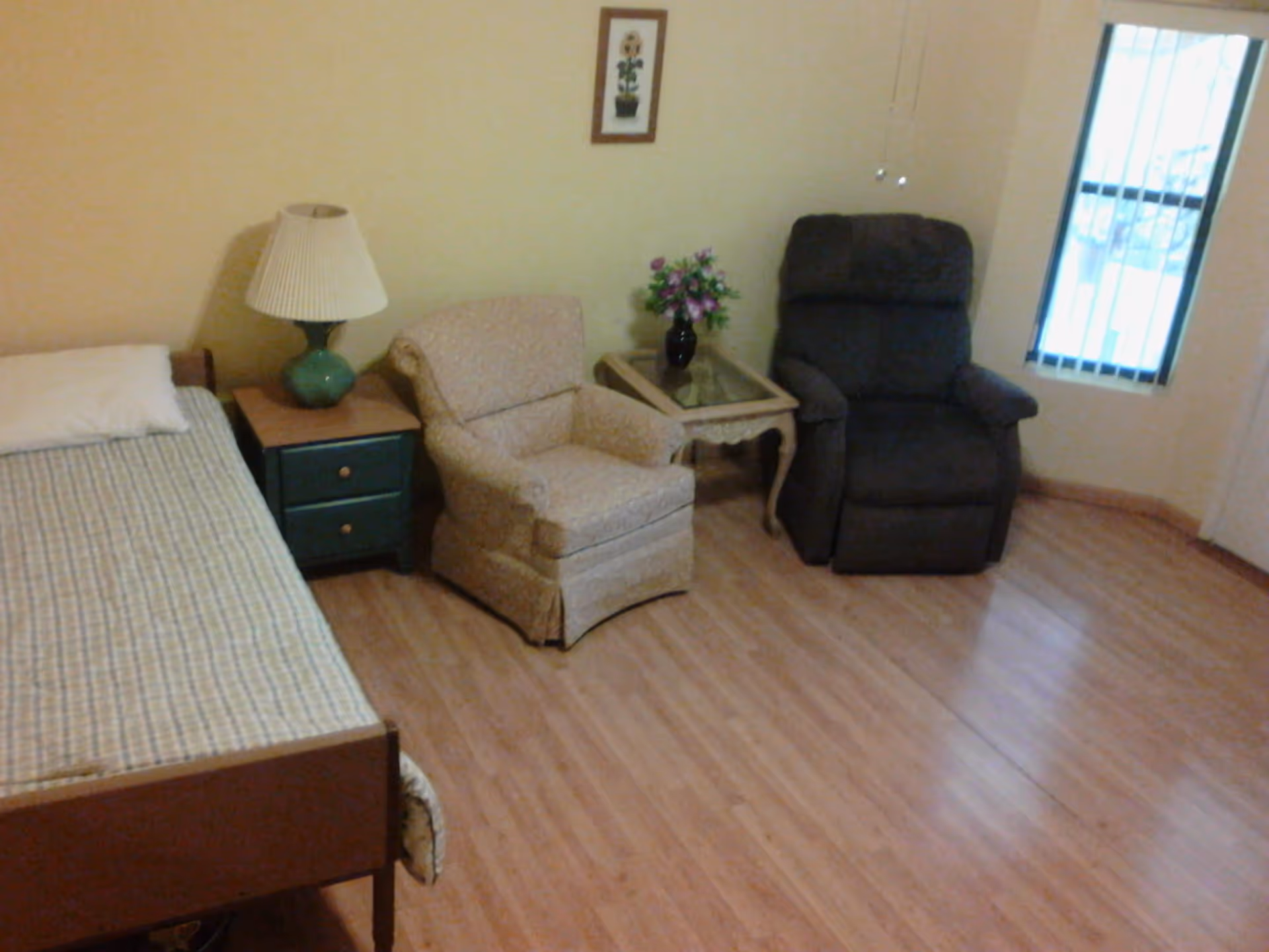 A simple bedroom with a single bed covered in a checkered bedspread, a green nightstand with a lamp, two armchairs, a small glass-top side table with a vase of flowers, a framed picture on the wall, and a window with vertical blinds.