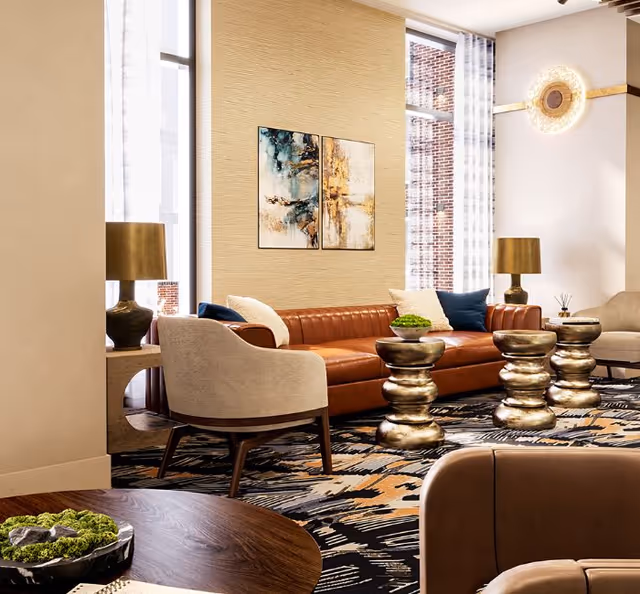 A modern living room with a brown leather sofa adorned with white and blue pillows, two beige armchairs, three metallic gold side tables, and a patterned carpet. The room features large windows with sheer curtains, two gold table lamps, abstract artwork on the wall, and a round wooden coffee table with a green plant centerpiece.