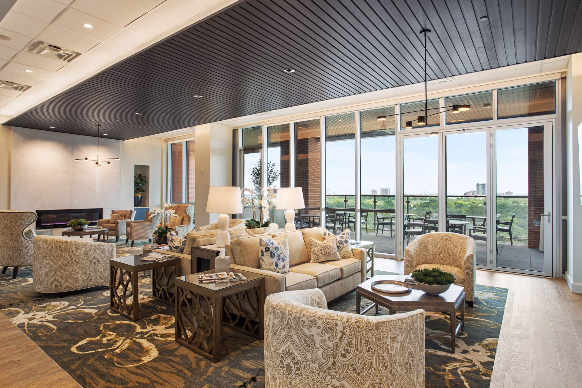 A spacious and well-lit living room area with comfortable beige sofas and patterned armchairs arranged around wooden coffee tables. The room features large floor-to-ceiling windows and glass doors that open to an outdoor patio with tables and chairs, offering a view of greenery and distant buildings. The ceiling is dark with recessed lighting, and the room is decorated with lamps, plants, and a patterned rug.