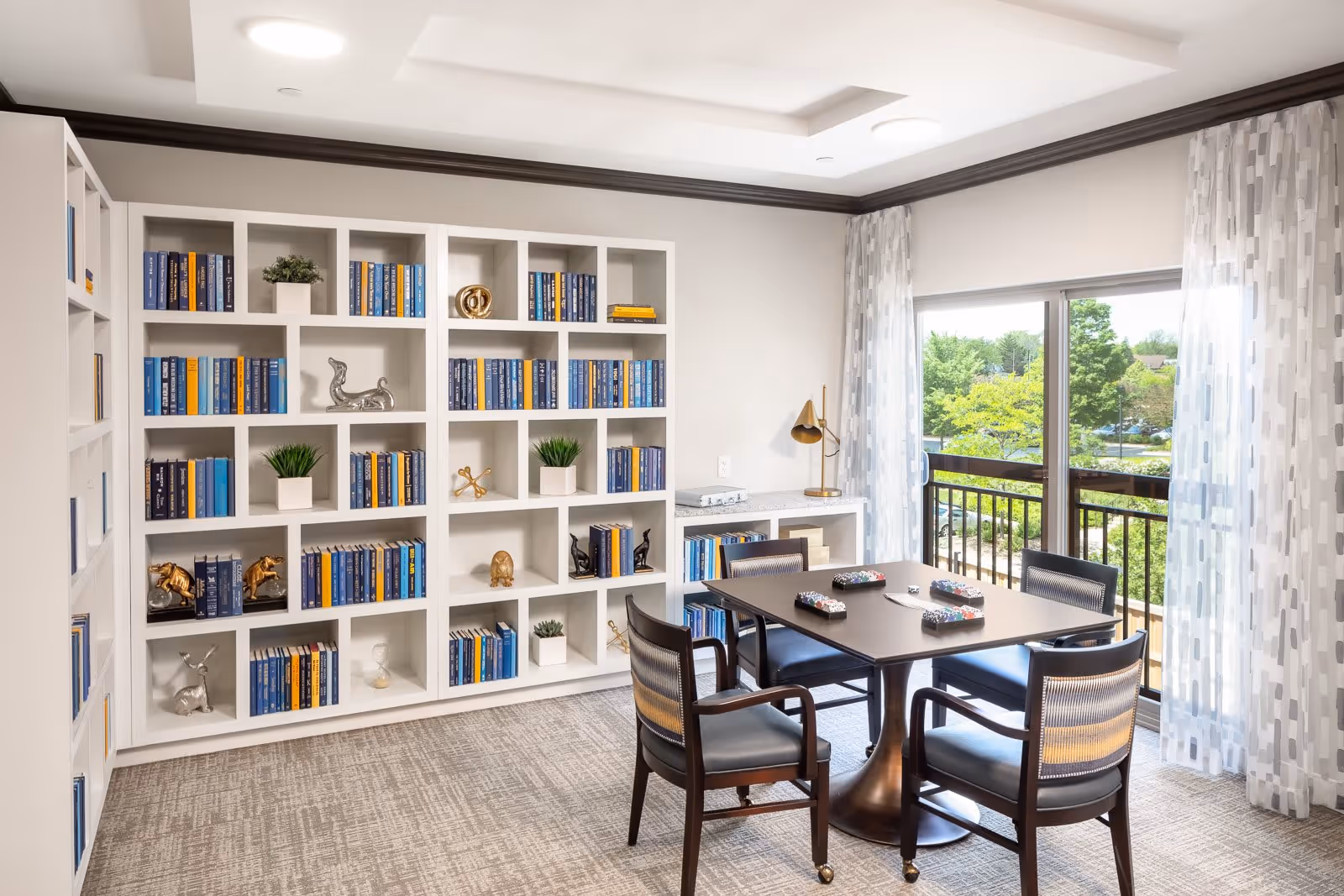 A bright room with a large white bookshelf filled with books and decorative items, a square table with four chairs around it, and a sliding glass door leading to a balcony with a view of trees and greenery outside. The room has light-colored walls, carpeted floor, and sheer patterned curtains.