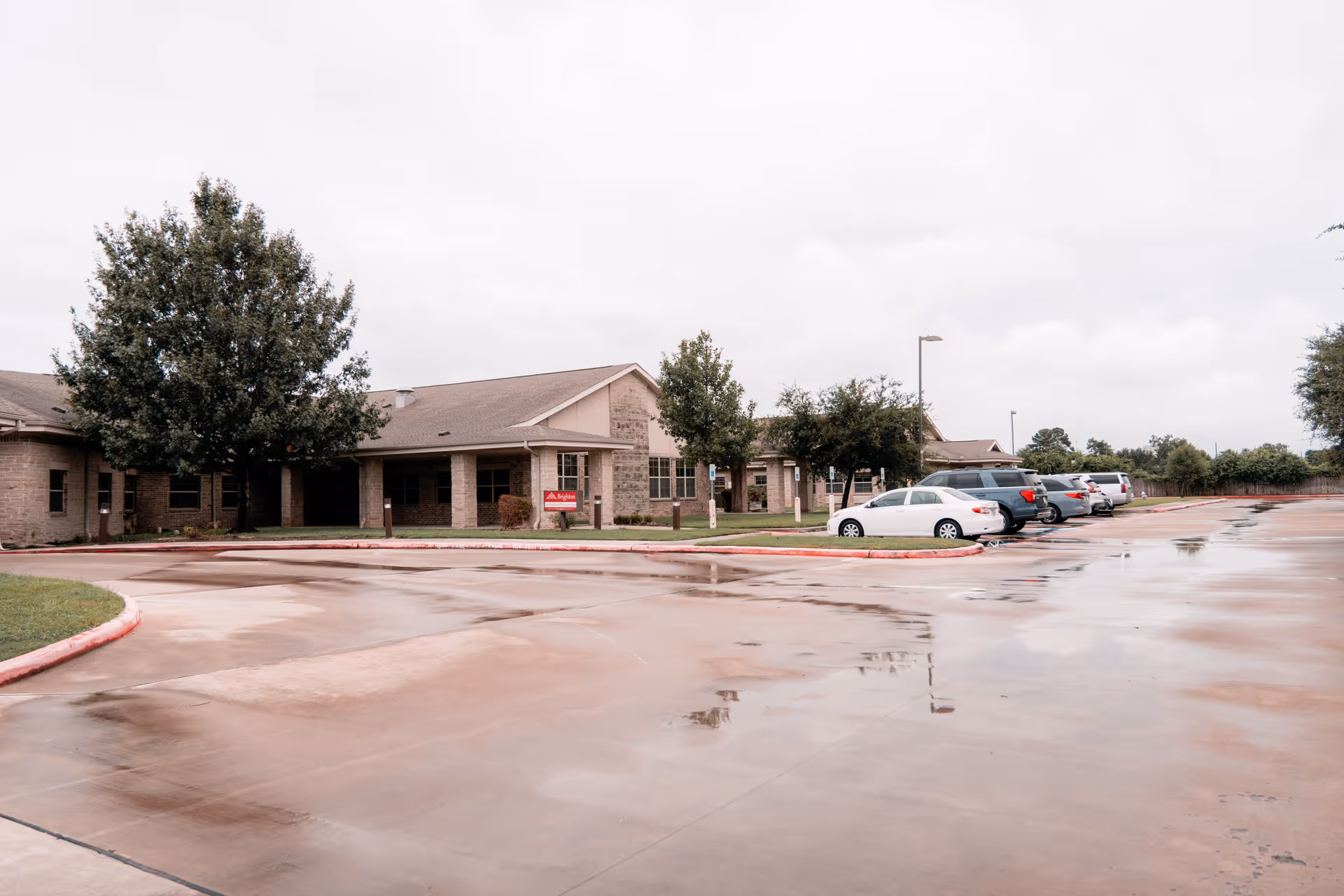 Exterior view of Brighton Senior Living facility showing a single-story building with a stone facade, several trees, a wet parking lot with multiple parked cars, and a cloudy sky.
