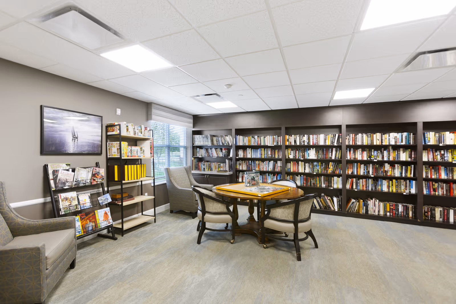 A well-lit library room with large bookshelves filled with books along the back wall. In the center, there is a round wooden table with four cushioned chairs around it. To the left, there are two armchairs, a magazine rack with various magazines, and a shelving unit with board games and yellow binders. A window with blinds lets in natural light.
