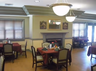 Dining room with several tables covered in red tablecloths, each set with chairs. The room features a fireplace with a mirror and framed pictures above it, windows with blinds, and hanging ceiling lights providing illumination.