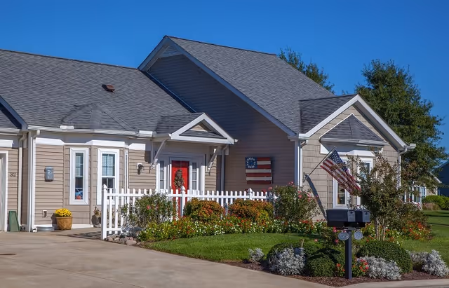 Exterior view of a single-story residential building with beige siding, a gray shingled roof, a white picket fence, and a red front door decorated with a wreath. There are bushes and flowers in the garden, an American flag and a Betsy Ross flag mounted on the wall, and a mailbox with two flags in front of the house under a clear blue sky.
