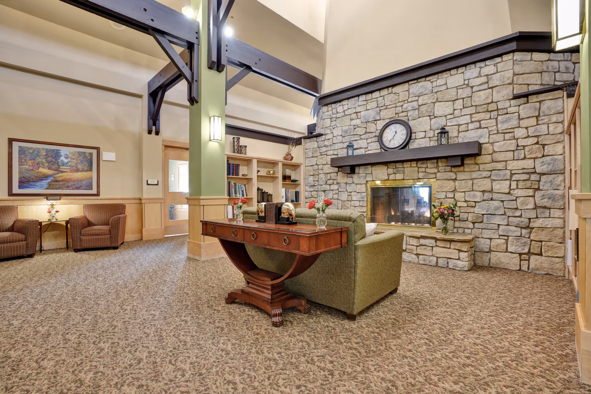 A cozy senior living facility common area with a stone fireplace, a clock above the mantel, and a green upholstered sofa facing the fireplace. There is a wooden table behind the sofa with decorative items and flowers. The room has high ceilings with exposed dark wooden beams, carpeted floor, built-in bookshelves, and additional seating with two armchairs near a framed landscape painting on the wall.