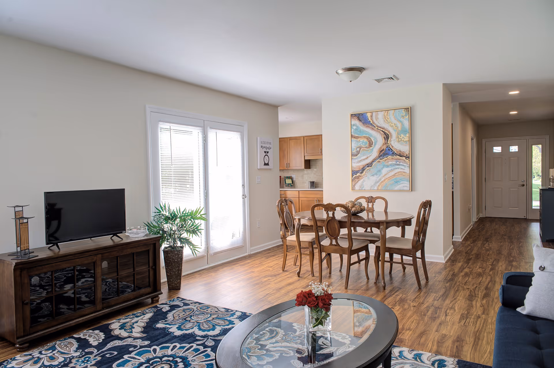 A bright and spacious living and dining area with wooden flooring. The living room features a dark wooden TV stand with a flat-screen TV, a potted plant, and a patterned blue and white rug. The dining area has a wooden dining table with six chairs and a colorful abstract painting on the wall. In the background, there is a kitchen with wooden cabinets and a hallway leading to the front door.