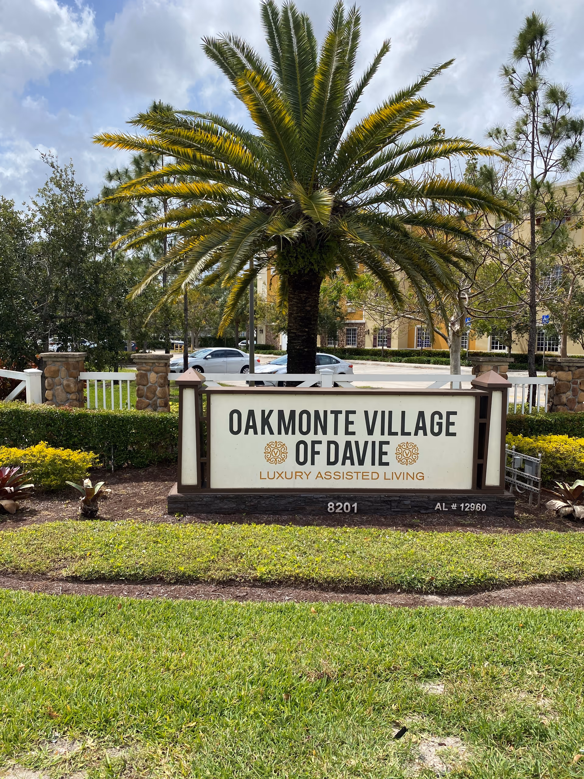 Entrance sign reading "Oakmonte Village of Davie Luxury Assisted Living" set in landscaped grounds with a large palm tree behind it.
