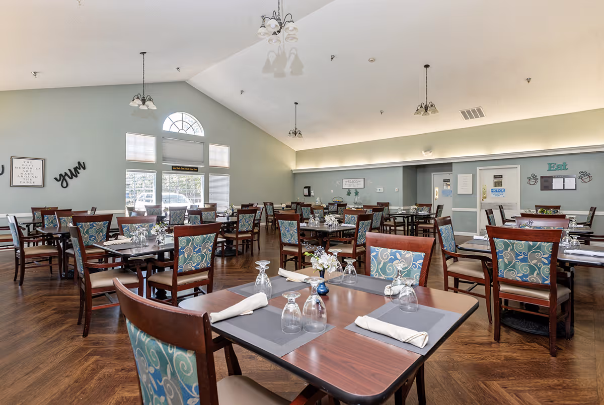 A spacious dining room with multiple wooden tables and chairs arranged neatly. Each table is set with placemats, napkins, and upside-down drinking glasses. The room has large windows allowing natural light to fill the space, light green walls, and several hanging light fixtures from a vaulted ceiling. Wall decorations include framed quotes and the words 'yum' and 'Eat'.