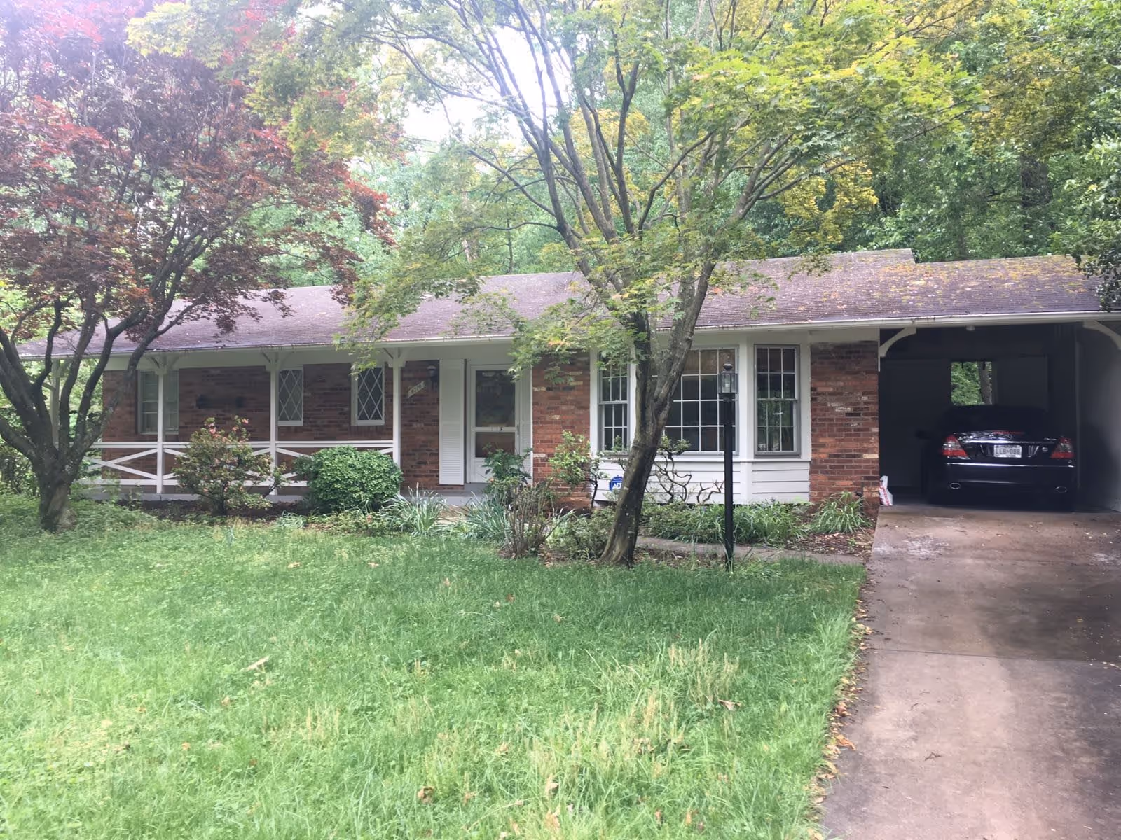 Front exterior view of a single-story brick house with a covered carport on the right side. The house has white framed windows and a white front door with a small porch area. There are several trees and bushes in the front yard with a green lawn.