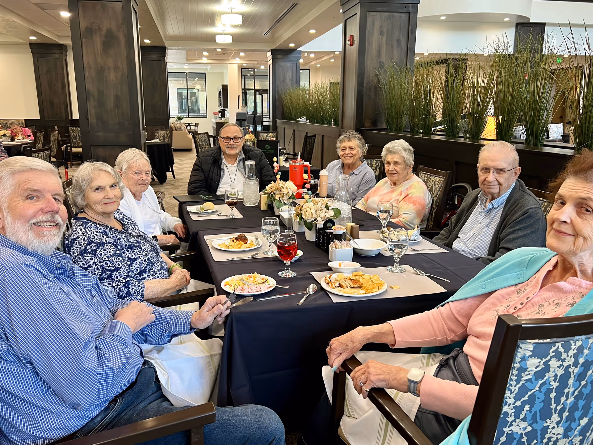 A group of eight elderly people sitting around a dining table in a senior living facility, enjoying a meal together. The table is set with plates of food, glasses of water and juice, and condiments. The room has dark wooden pillars and decorative plants, with a bright and spacious interior.