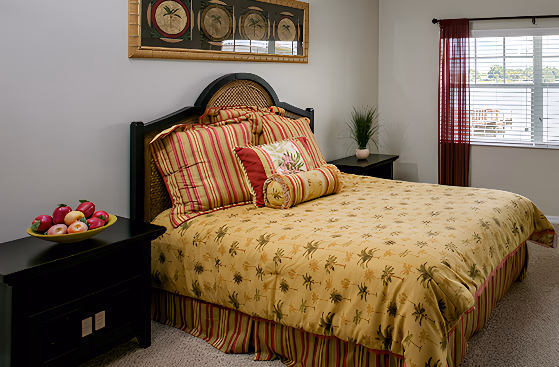 Bright bedroom with a large bed dressed in yellow floral bedding and striped pillows, two dark bedside tables (one with a bowl of fruit), wall art above the headboard, and a window with blinds.