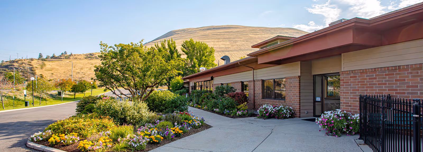 Exterior view of Riverside Health & Rehabilitation facility with a brick building, a concrete walkway, and landscaped flower beds with colorful flowers and green shrubs. There is a tree near the walkway and a hill in the background under a partly cloudy sky.