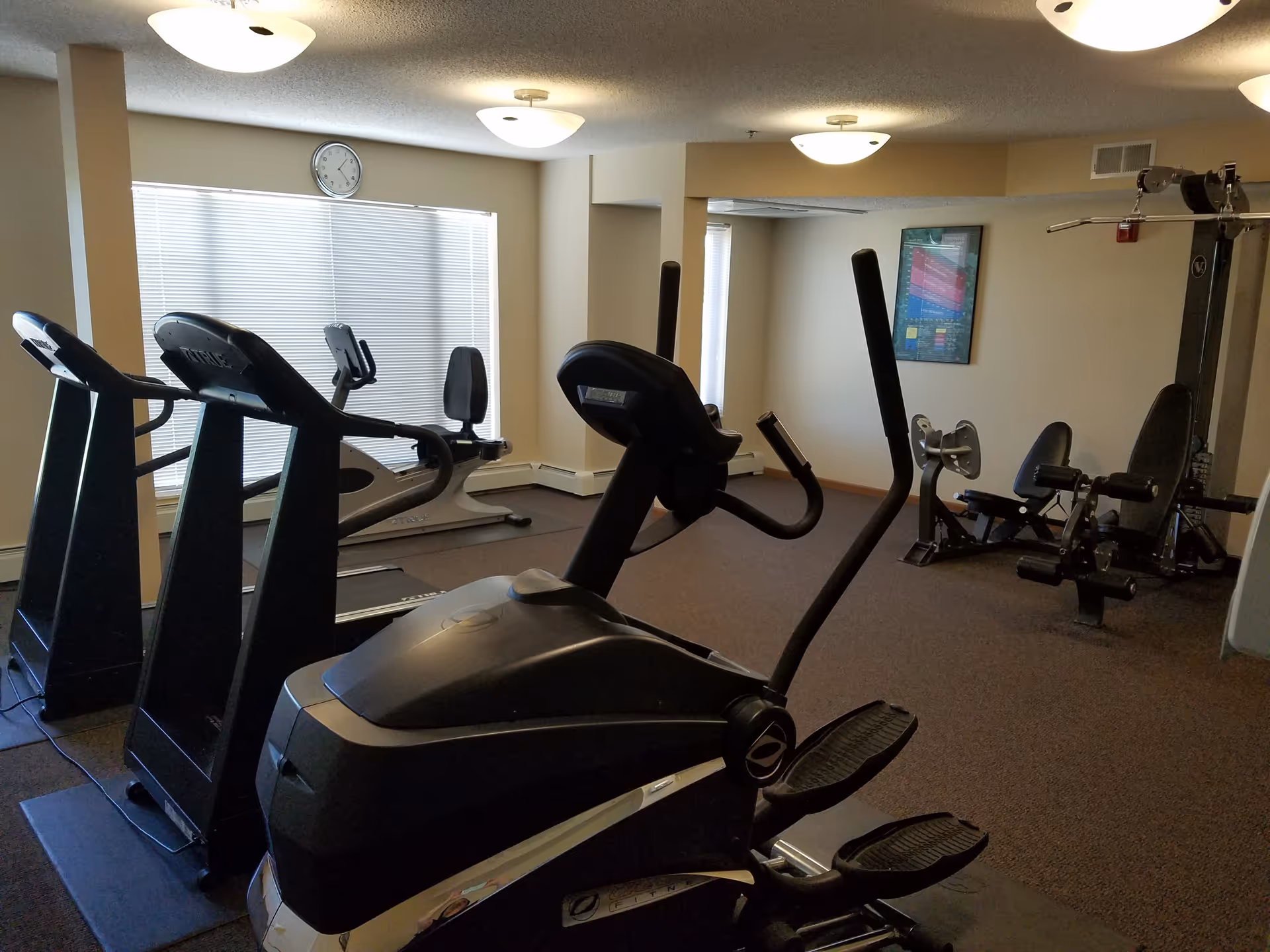 Interior view of a fitness room with exercise equipment including treadmills, an elliptical machine, and weight machines. The room has beige walls, carpeted floor, ceiling lights, a clock on the wall, and a window with blinds.