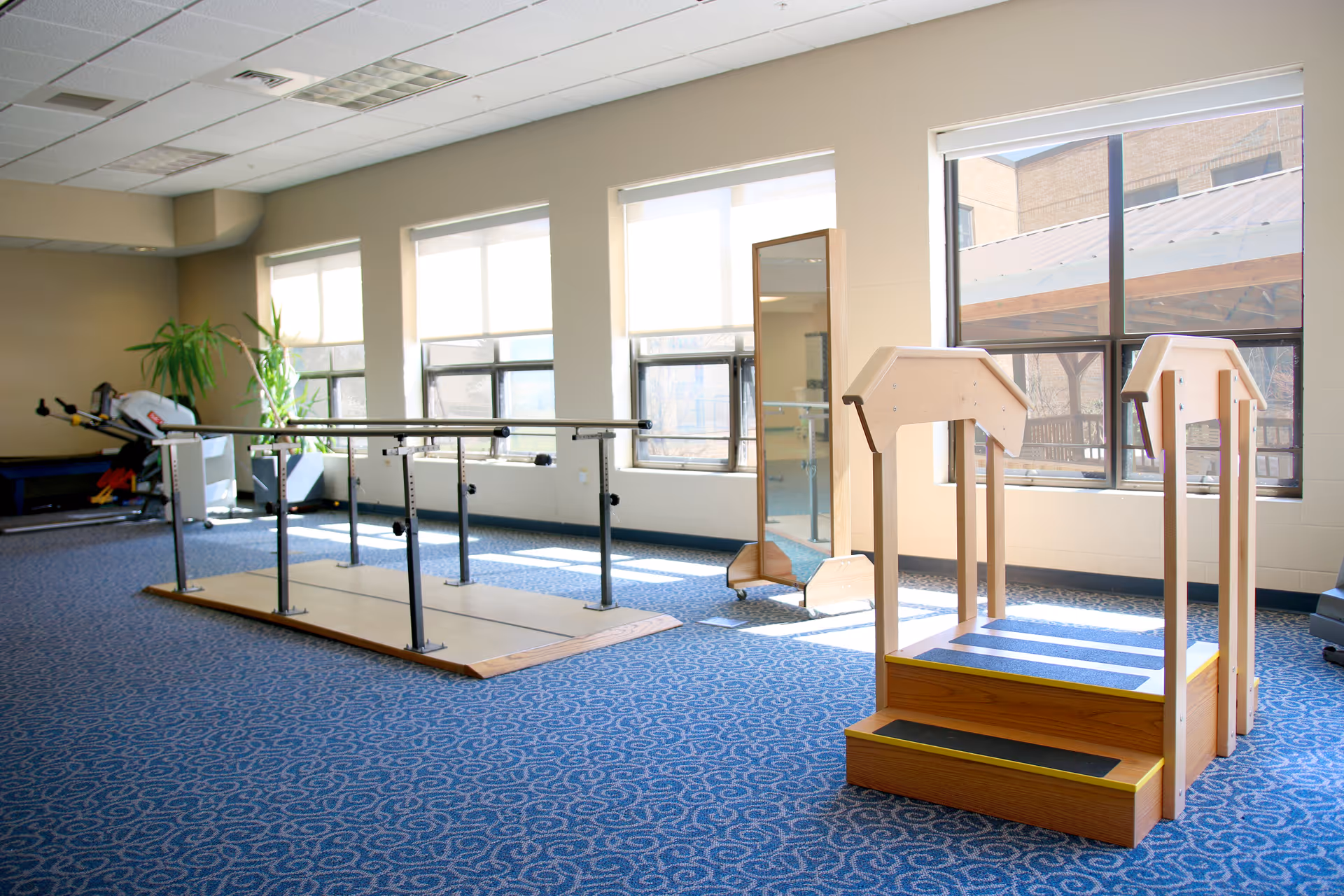 A rehabilitation room with parallel bars for walking exercises, a small wooden staircase with handrails, large windows letting in natural light, a full-length mirror, and exercise equipment in the background.