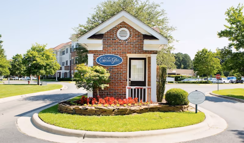 Small brick guardhouse with a sign reading 'Cypress Glen' surrounded by a landscaped flower bed and greenery, located at the entrance of a senior living community with multi-story residential buildings and trees in the background.