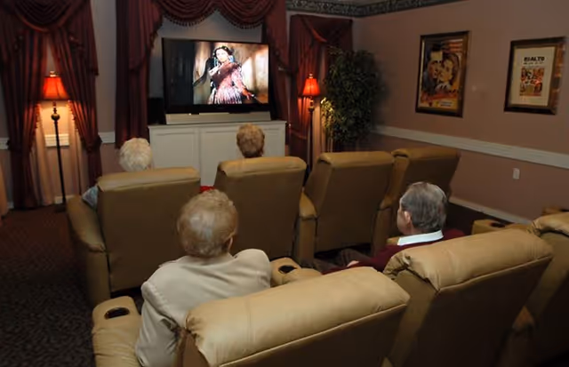 A cozy movie room with four elderly people seated in beige recliners facing a large flat-screen TV playing a classic film. The room is warmly lit with two floor lamps and decorated with framed vintage movie posters and burgundy curtains.