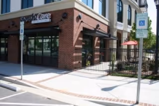 Exterior view of SarahCare at Lake Boone Trail building entrance with brick and beige walls, large windows, a black metal fence, and a sidewalk with handicap parking signs.