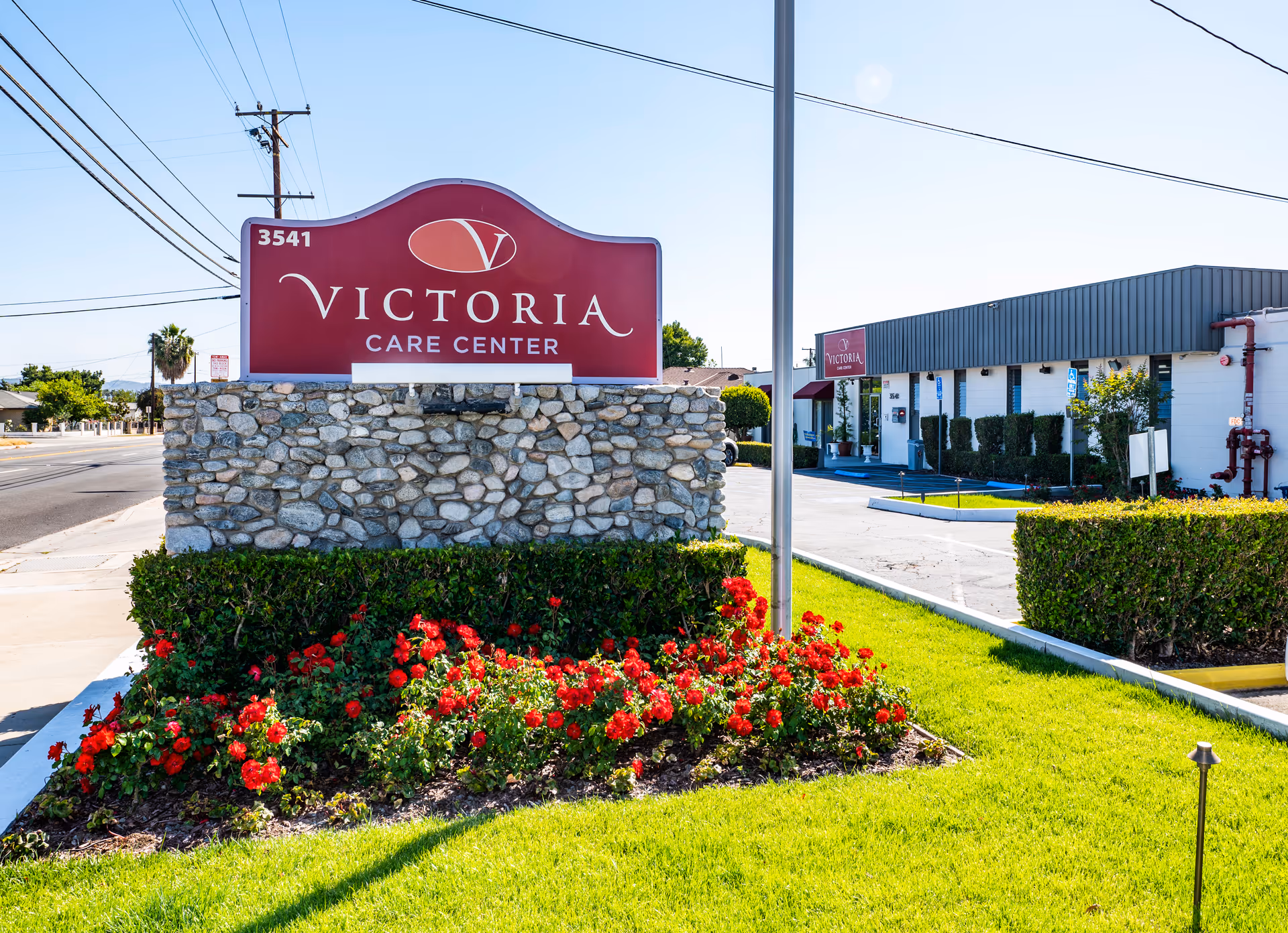 A stone sign reading "Victoria Care Center" stands amid red flowers in front of the care facility and parking lot.