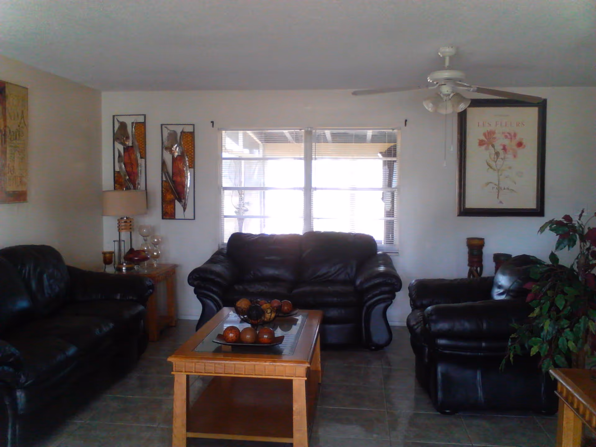 A living room with three black leather sofas arranged around a wooden coffee table with decorative items on it. The room has a tiled floor, a ceiling fan with lights, wall art including a framed floral print and metal sculptures, a table lamp, and a potted plant.