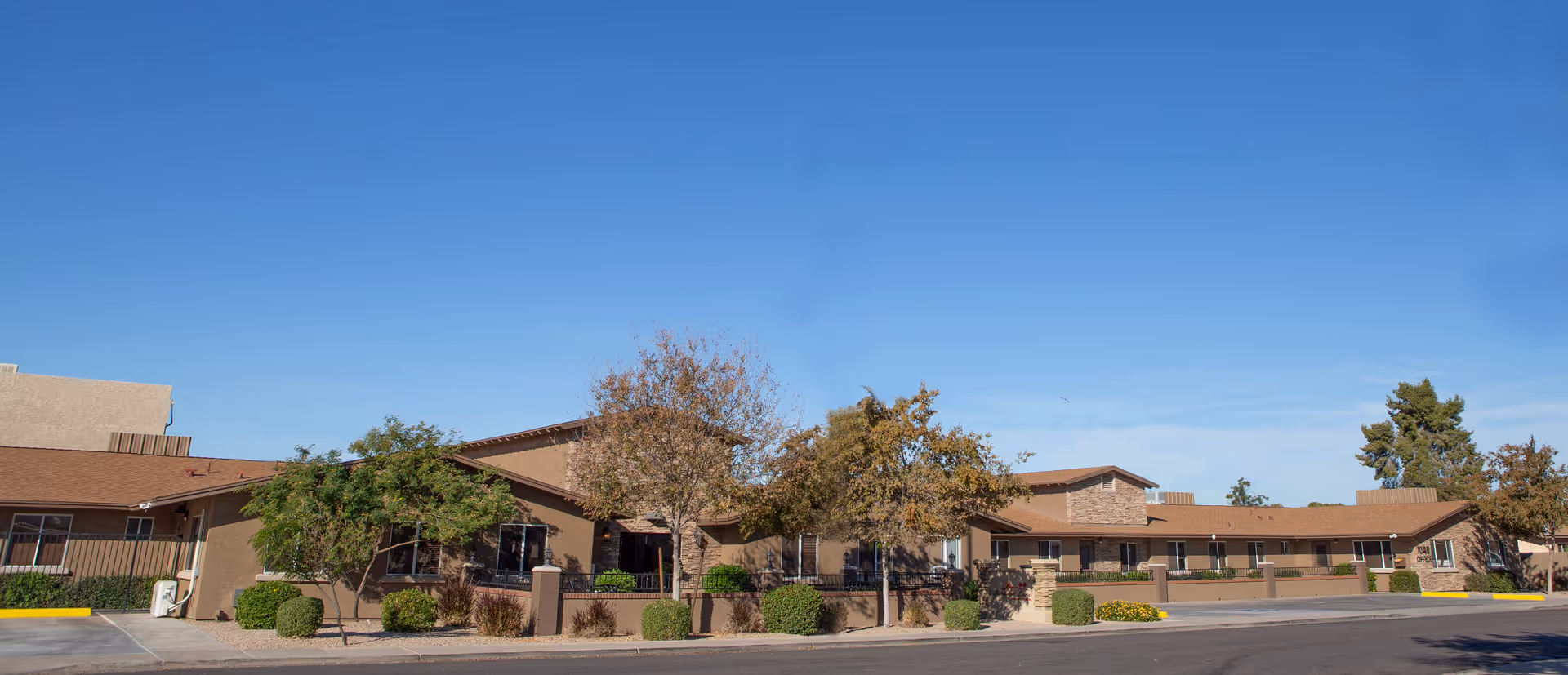 Exterior view of a single-story building with a brown roof and beige walls, surrounded by trees and shrubs under a clear blue sky. The building has multiple windows and a sign near the entrance that reads '1040 OFFICE'.