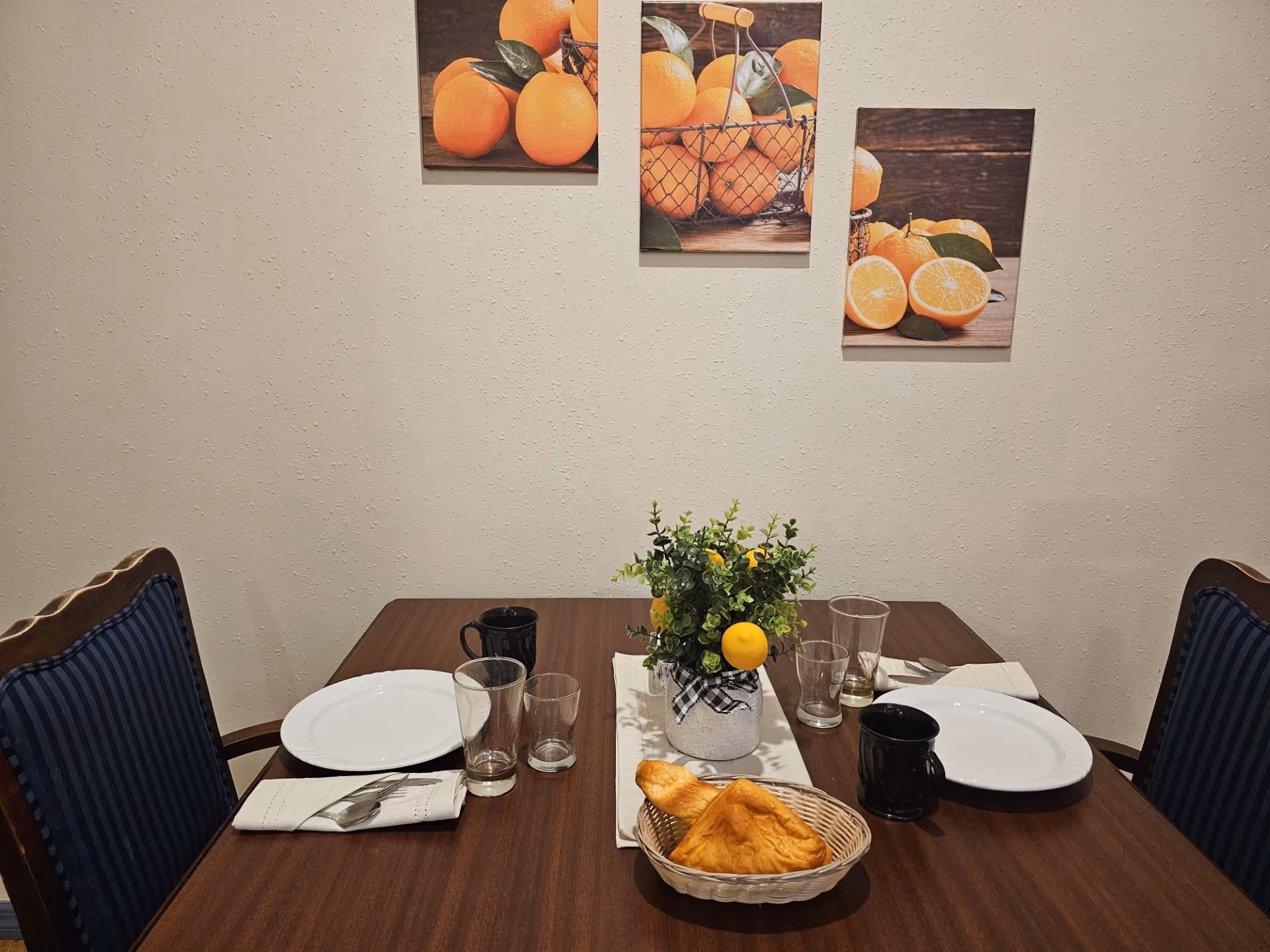 Dining table set for two with plates, glasses, mugs, a bread basket and a small floral centerpiece beneath orange-themed wall art.