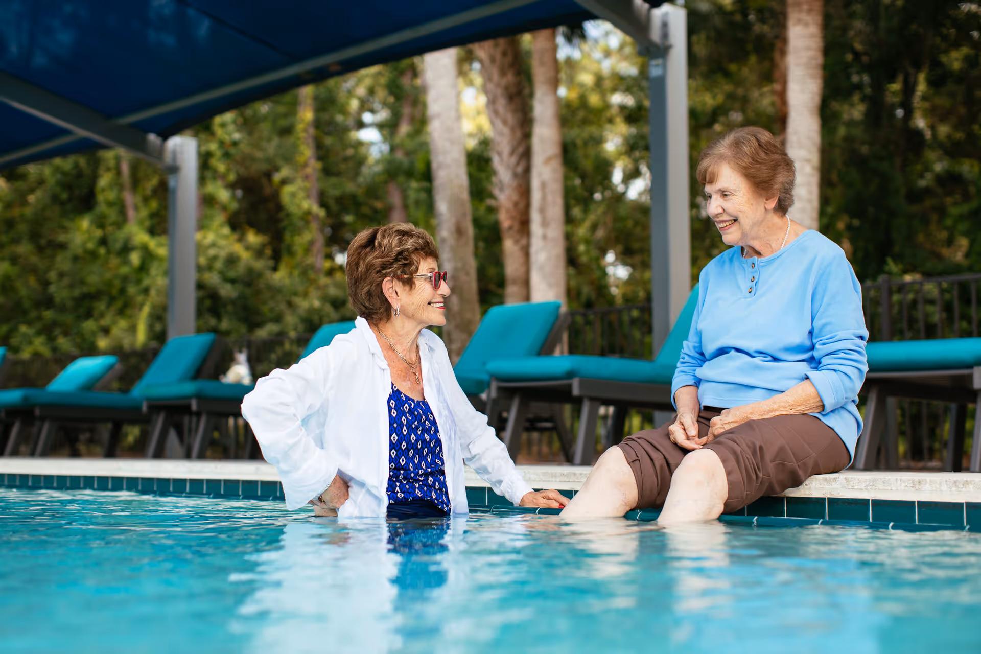 Two elderly women enjoying time by a swimming pool. One woman is sitting on the edge of the pool with her feet in the water, wearing a blue long-sleeve shirt and brown pants. The other woman is standing in the pool, wearing sunglasses, a white jacket, and a blue patterned top. There are lounge chairs with teal cushions and trees in the background.